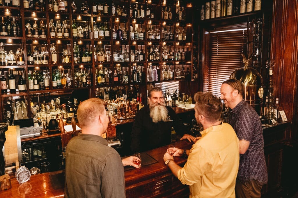 Three patrons are ordering drinks at the Two Fingers Gentleman's Bar. A large selection of whiskey bottles and other drinks are on the wall behind the bartender.